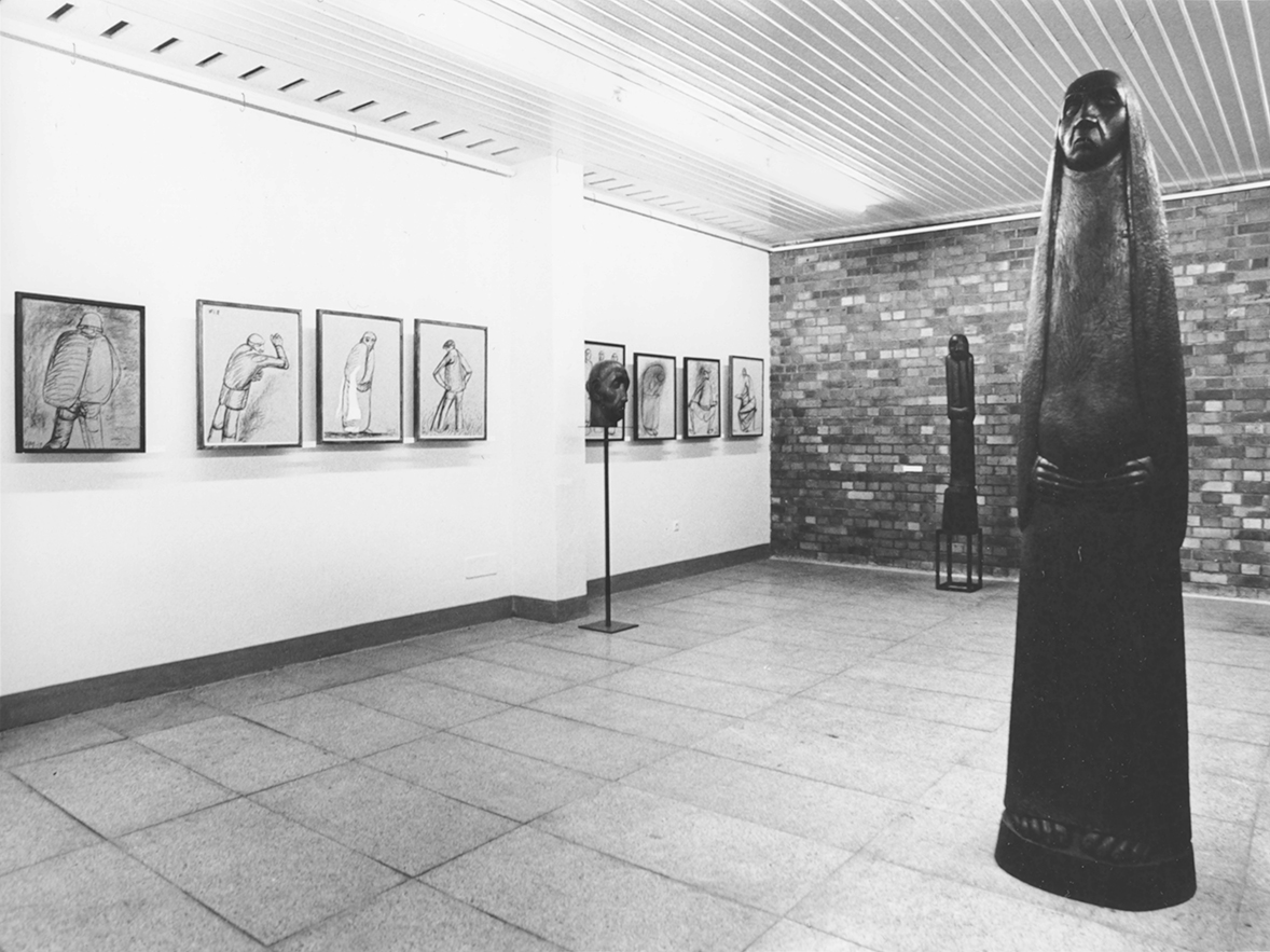 zur Vergrößerungsansicht des Bildes: Schwarz-weiß-Foto mit Blick in einen Galerie Raum, links an der Wand hängen Zeichnungen und Grafiken, rechts im Raum steht im Vordergrund eine hohe, schmale Skulptur, die eine Frau darstellt 