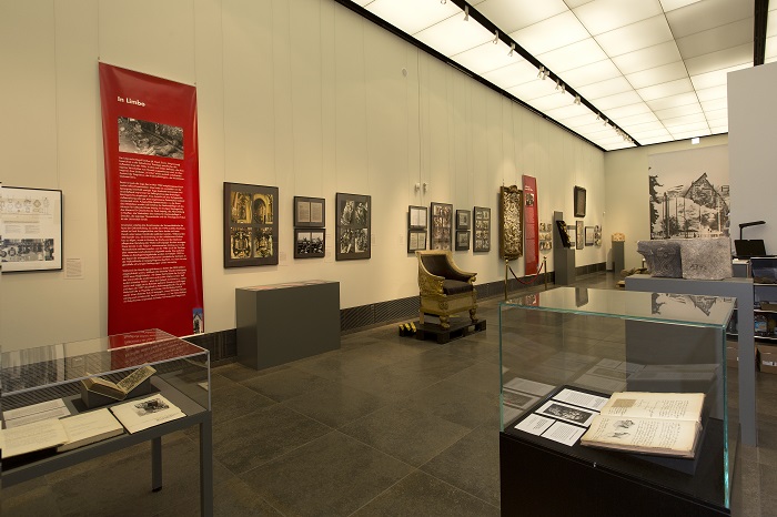 Blick in den Galerieraum mit Vitrinen und Objekten, an den Wänden hängen Textfahnen und Fotos