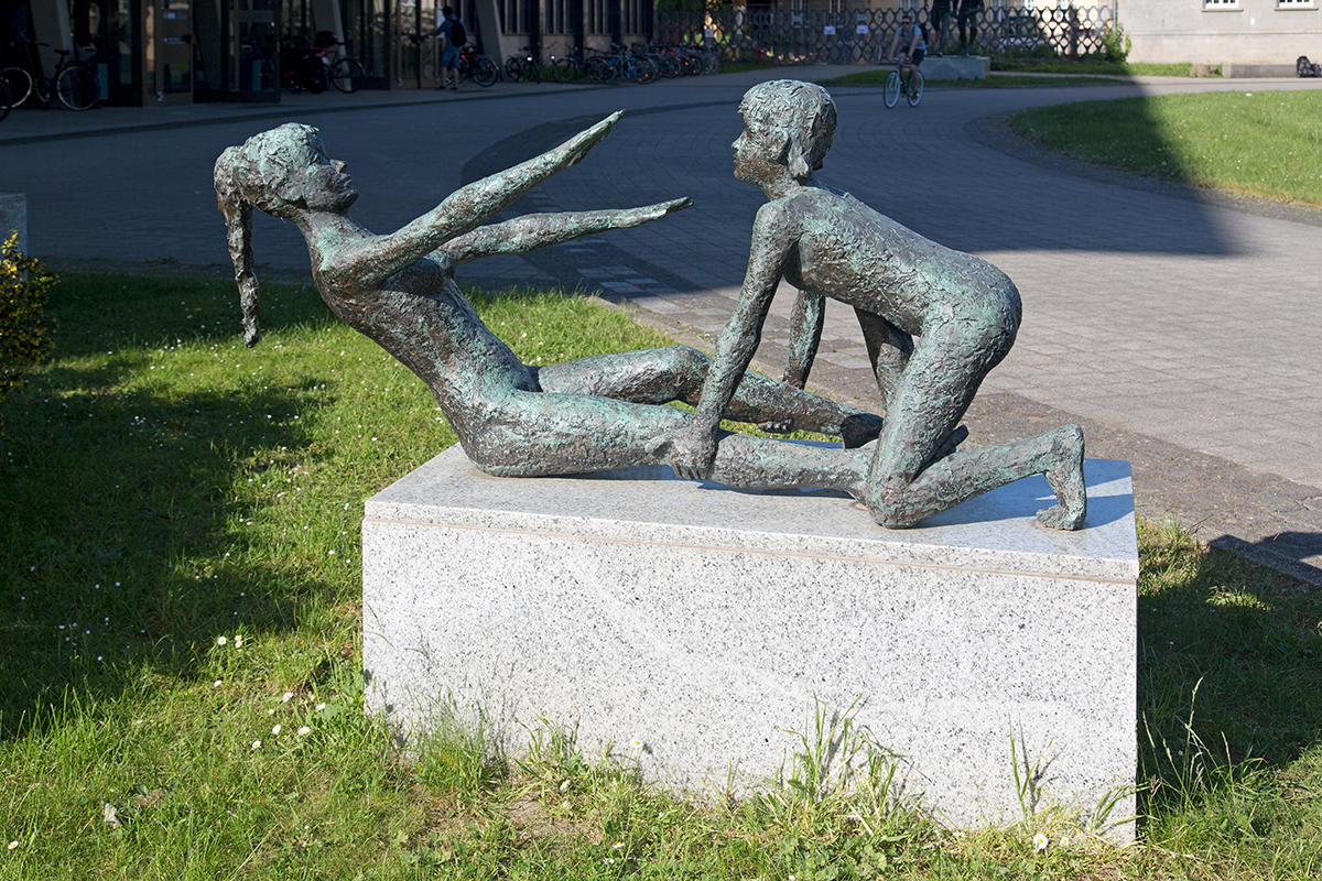 zur Vergrößerungsansicht des Bildes: Bronzeskulptur auf dem Campus Jahnallee, zwei Figuren aus Bronze auf einem Steinsockel, ein Mädchen und ein Junge bei Turnübungen, das Mädchen führt die Übung "Rumpfheben" aus, der Junge hält ihre Beine zur Unterstützung fest