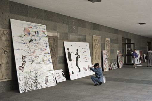 An einer Wand des Universitätsgebäudes lehnen mehrere Tafeln mit Pochoir-Kunstwerken, davor sitzt eine Künstlerin und betrachtet ihr Kunstwerk