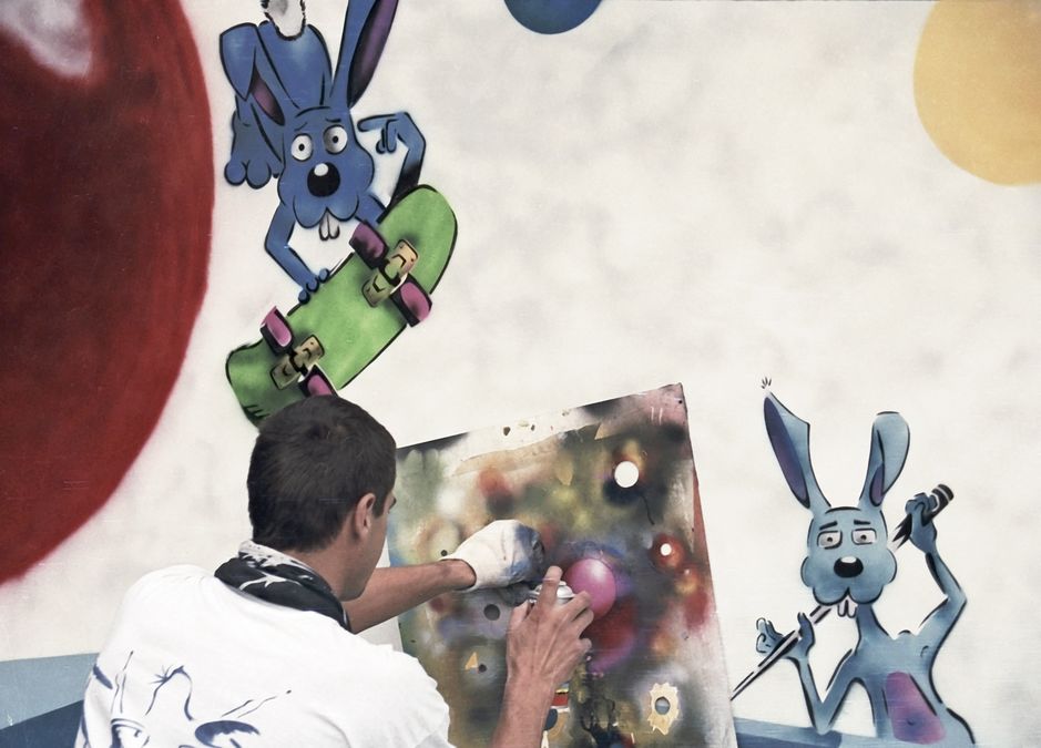 zur Vergrößerungsansicht des Bildes: Vor einer Wand mit Graffiti (zwei blaue Hasen, einer davon fährt Skateboard) sieht man einen Graffiti-Künstler bei der Arbeit, er sprüht gerade mit einer Dose Farbe auf eine Tafel, auf der abstrakte bunte Formen zu sehen sind
