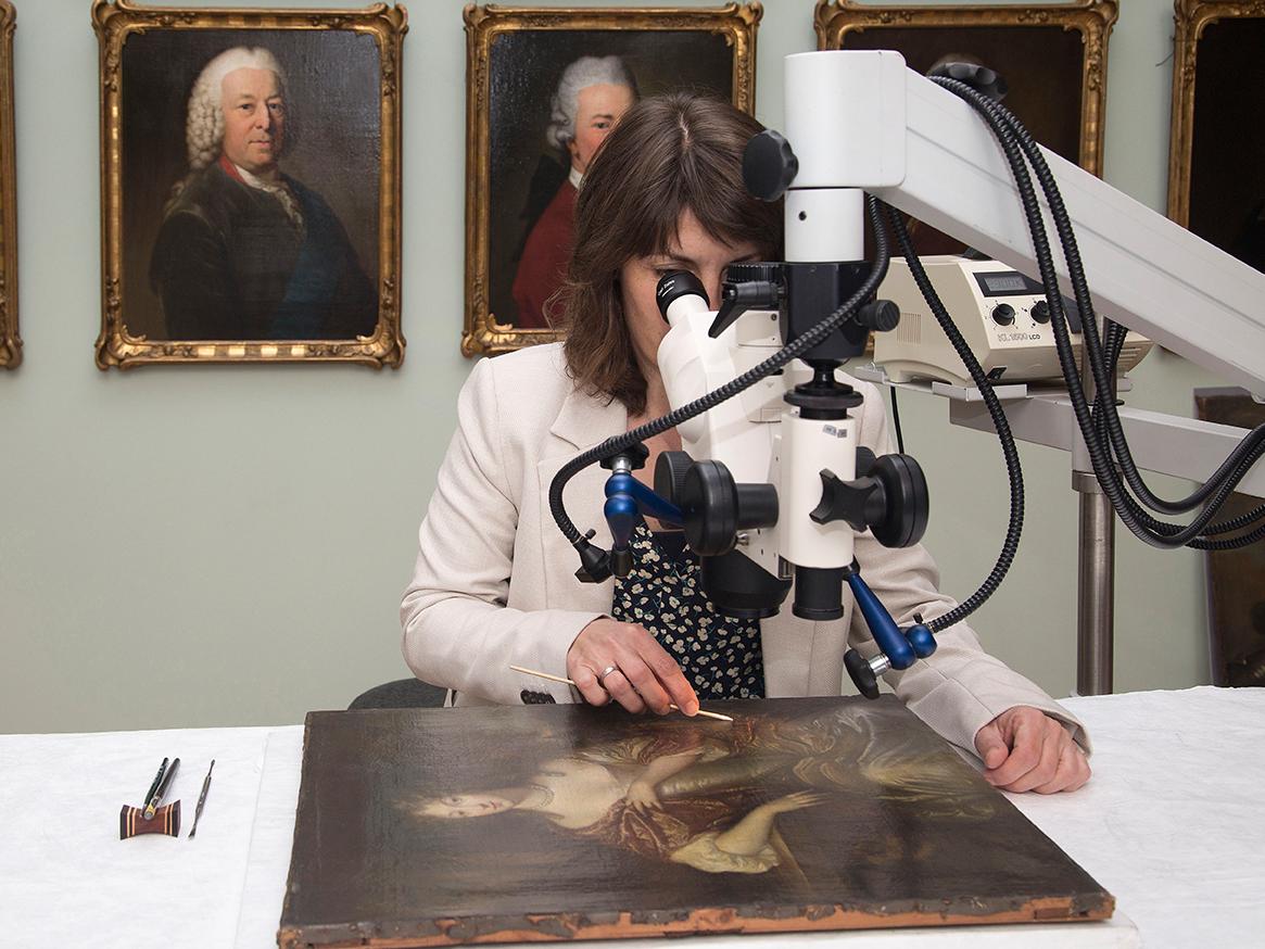 zur Vergrößerungsansicht des Bildes: Eine Restauratorin prüft mit einem Mikroskop ein Gemälde.