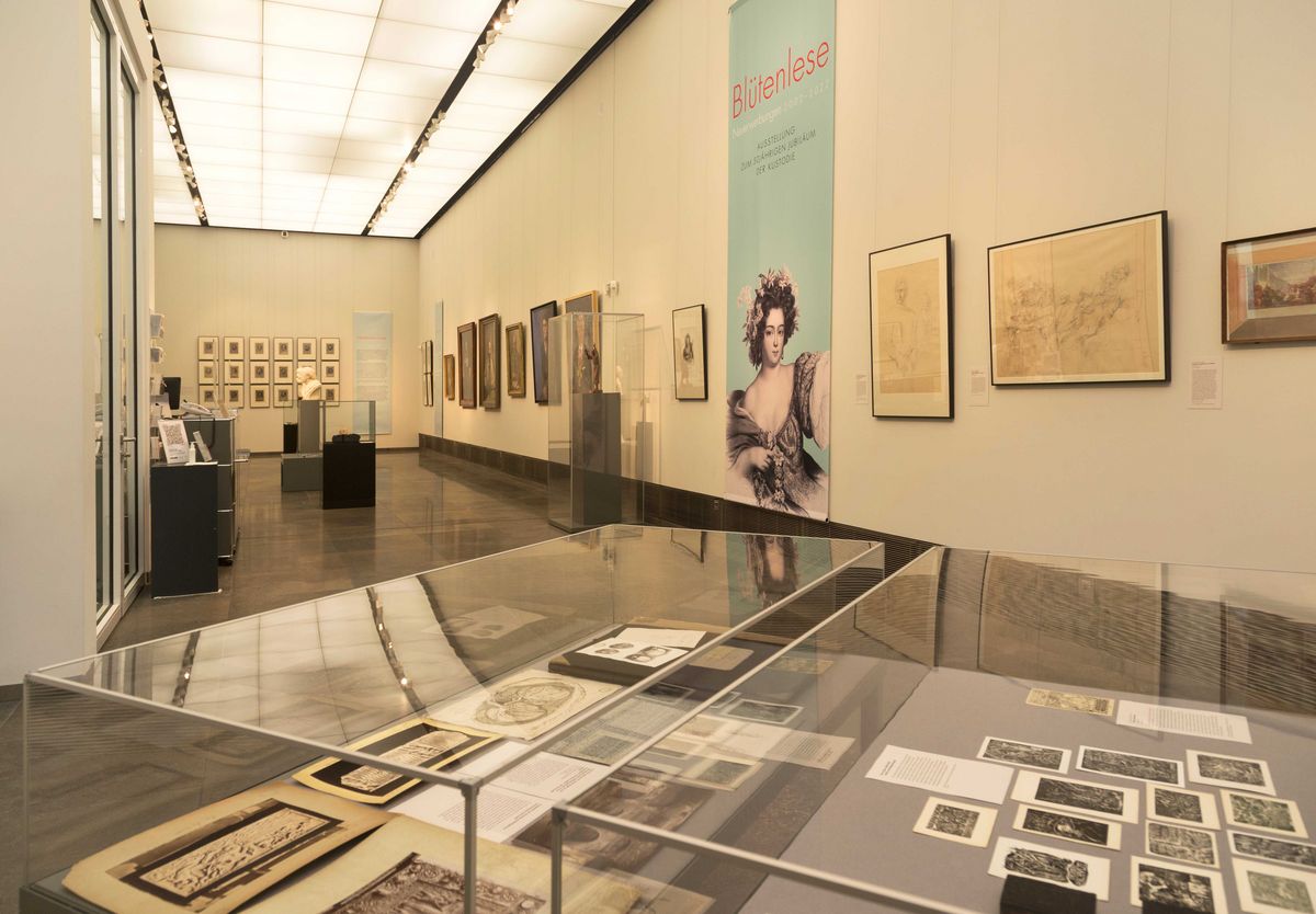 zur Vergrößerungsansicht des Bildes: Blick in den Galerieraum, Neues Augusteum, mit der Ausstellung "Blütenlese". Im Vordergrund sind Vitrinen mit Grafiken zu sehen, rechts an der Wand sind Zeichnungen von Werner Tübke, dahinter, der Tür gegenüber, eine große Fahne mit dem Ausstellungsmotiv, im Hintergrund ein Blick in den Raum mit Ölgemälden und Büsten