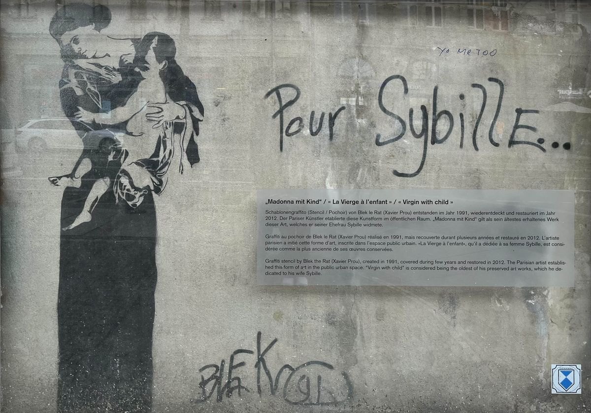 zur Vergrößerungsansicht des Bildes: Schablonen-Graffiti, Madonna mit Kind auf einer Häuserwand hnter Glast, ganz unten rechts das Denkmal-Zeichen, rechts daneben der Schriftzug "Pour Sybille ...", darunter eine Erläuterungstafel