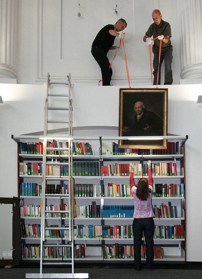 Drei Mitarbeiter der Kustodie bringen gemeinsam ein Gemälde in der Universitätsbibliothek Leipzig an.