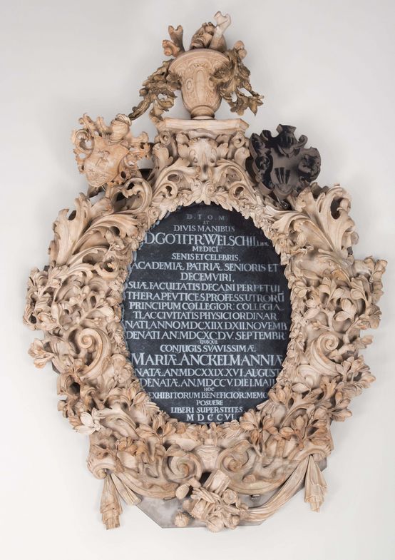 Farbfotografie: Detailaufnahme eines Gedächtnismal aus Alabaster mit kunstvollen Verzierungen und Ornamenten am Rand und einer schwarzen, ovalen Tafel in der Mitte mit weißer Schrift in lateinischer Sprache