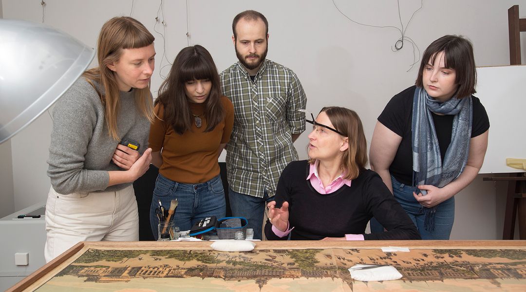 An einem Tisch vor einer Grafik mit einer Stadtansicht Leipzigs aus dem 19. Jahrhundert sitzt eine Papierrestauratorin. Um sie herum sind vier Studierende versammelt, drei Frauen und ein Mann. Sie lauschen den Erklärungen der Restauratorin. 