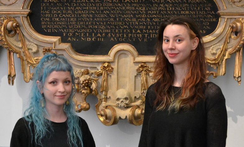 zwei junge Frauen, die die Führung im Rahmen des Wave Gotik Treffen anbieten, stehen vor einem Schnitzepitaph, direkt hinter ihnen ein geschnitzter Totenkopf, das Epitaph ist Weiß und Gold gefassttt Nicola Sommer und Alicia Opitz vor dem Epitaph für Heinrich Heideck, Foto: Sommer/Opitz