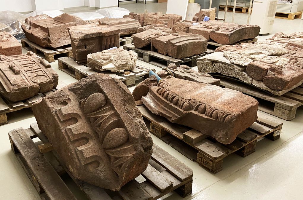 zur Vergrößerungsansicht des Bildes: Blick in den Kunstdepot, die Steinfragmente aus rotem Rochlitzer Porphyr liegen einzeln ausgebreitet auf Holzpaletten, im Vordergrund ist ein Teil mit Ornamenten, Eierstab, etc. zu sehen, dahinter Fragmente mit Wappen und Porträts