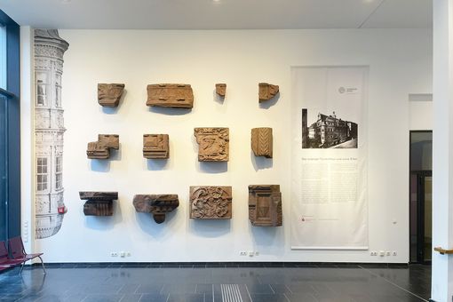 An der Wand sind zwölf Steinfragmente zu sehen (4 Steine in 3 Reihen übereinander), rechts ist eine Schrifttafel mit einem historischen Foto angebracht, darunter ein erklärender Text; links neben den Steinen ein Teil des Erkers fast über die ganze Wand, angeschnitten